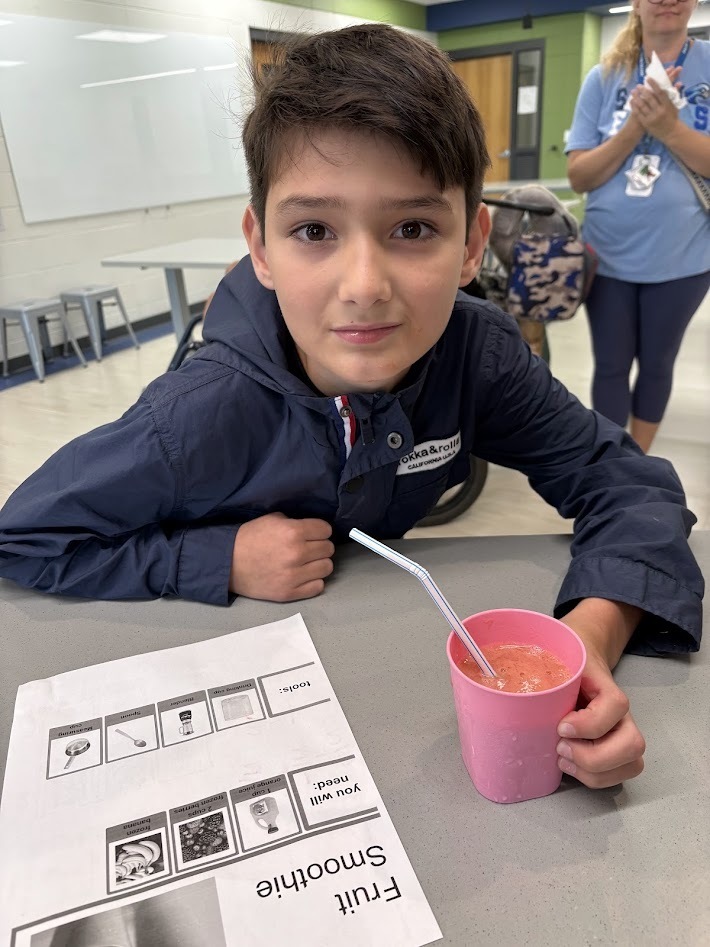 A boy sitting at a desk with a pink cup containing a smoothie and a recipe sheet titled "Fruit Smoothie". (