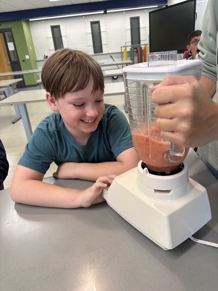 A smiling young boy in a blue shirt watching a blender make a pink fruit smoothie in a classroom setting.
