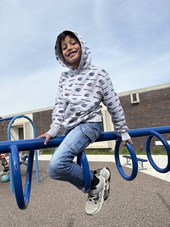 A smiling boy wearing a hooded sweatshirt sitting on a blue monkey bar structure at a playground.