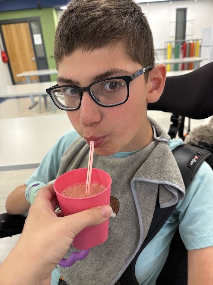 A student wearing glasses drinking a smoothie through a straw from a pink cup held by a teacher. (