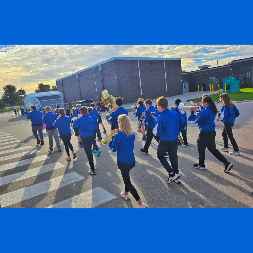 School marching band students in matching blue jackets walking and playing instruments along a road near school buildings.