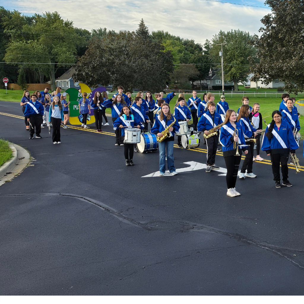 Members of a school marching band lined up with their instruments on an asphalt driveway near an athletic field