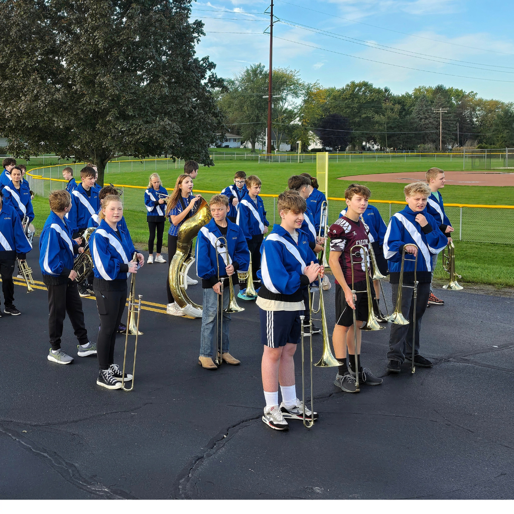 Members of a school marching band lined up with their instruments on an asphalt driveway near an athletic field