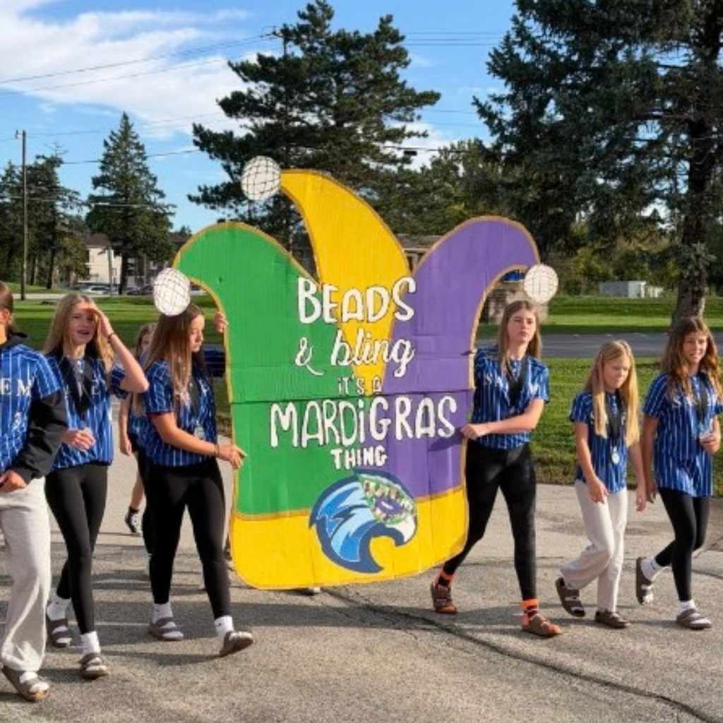 Students in matching blue striped shirts walking outside holding a large Mardi Gras jester hat sign.