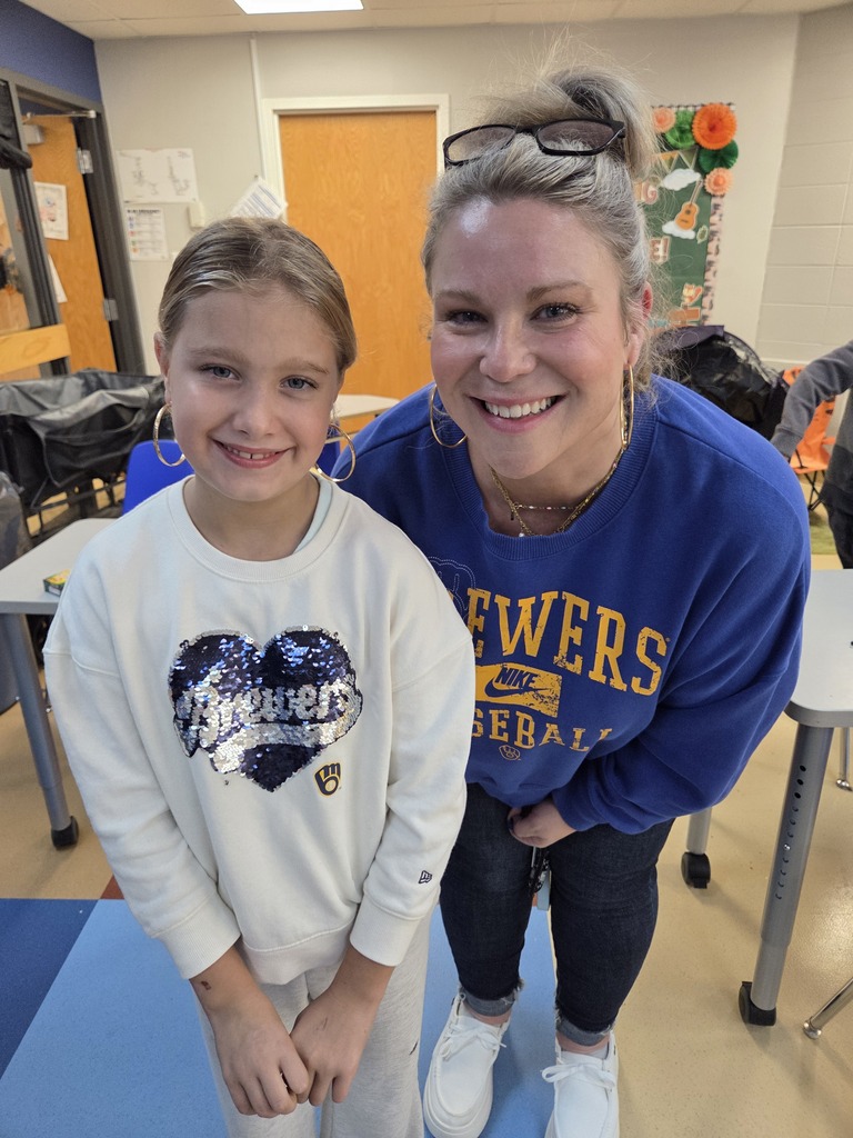 A smiling young student and teacher pose together in a classroom, both wearing hoop earrings and Milwaukee Brewers apparel.