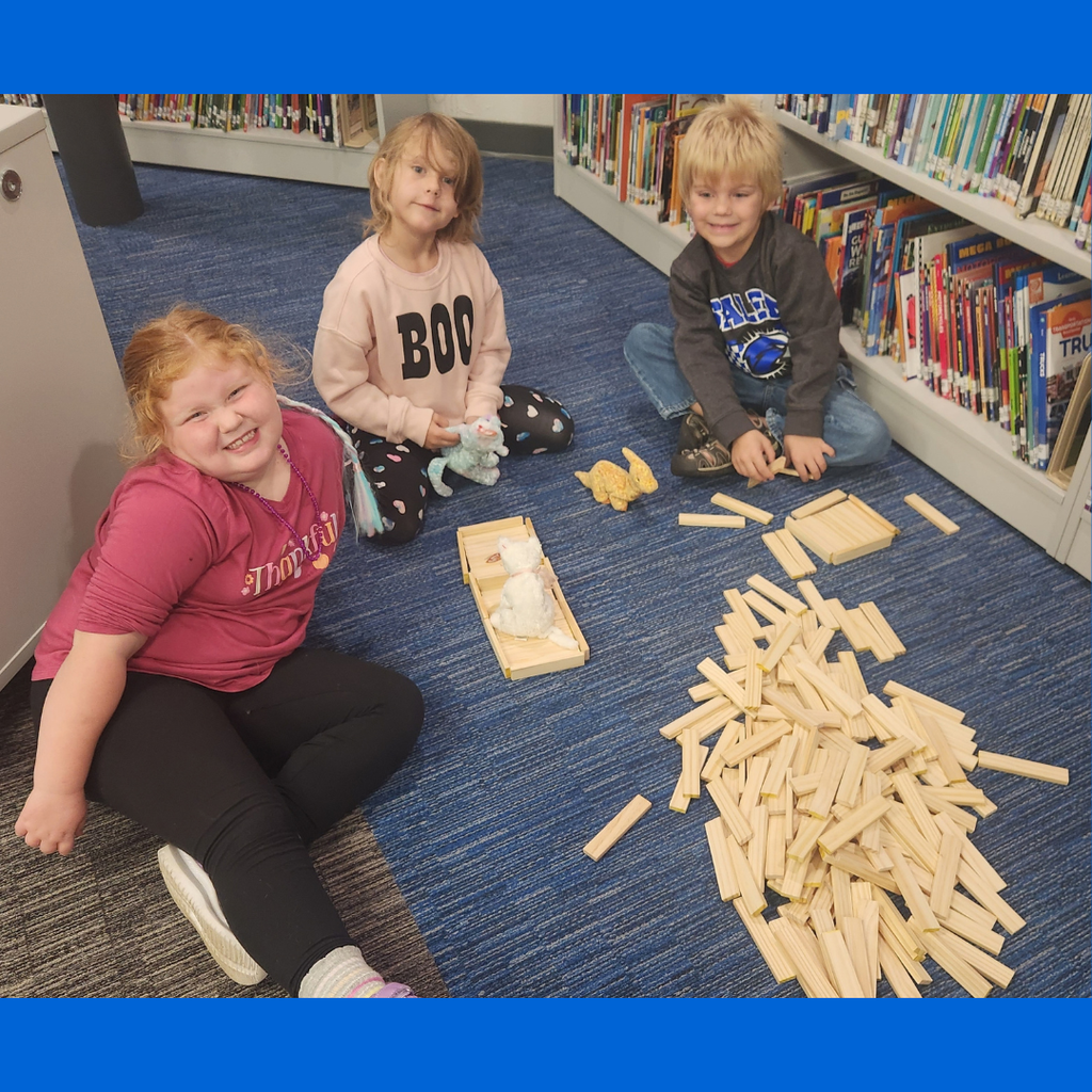 Three young students sit on the carpet in a school library, playing together with wooden building planks.