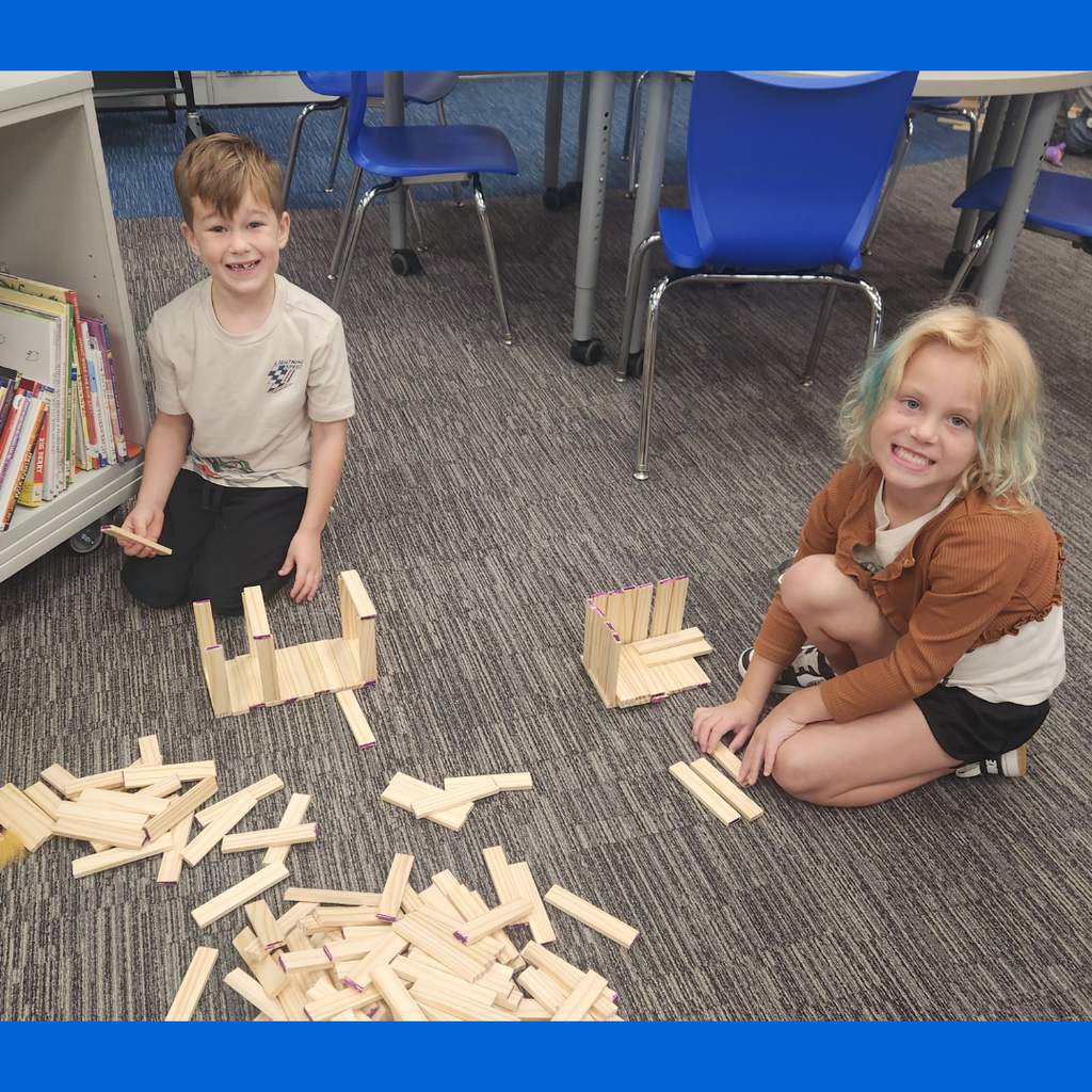 A boy and girl smile near bookshelves while assembling small structures on the floor using wooden planks.