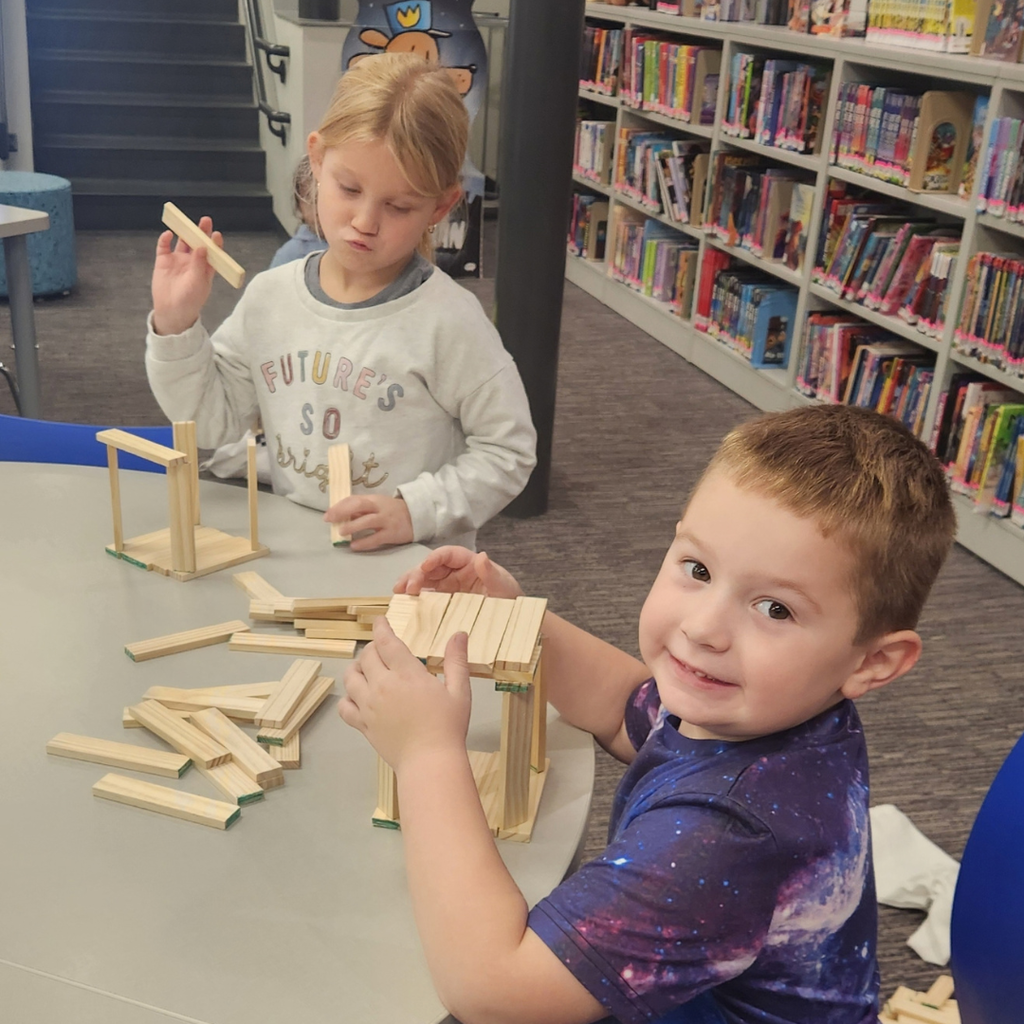 Two young students build wooden plank structures at a table in a school library.