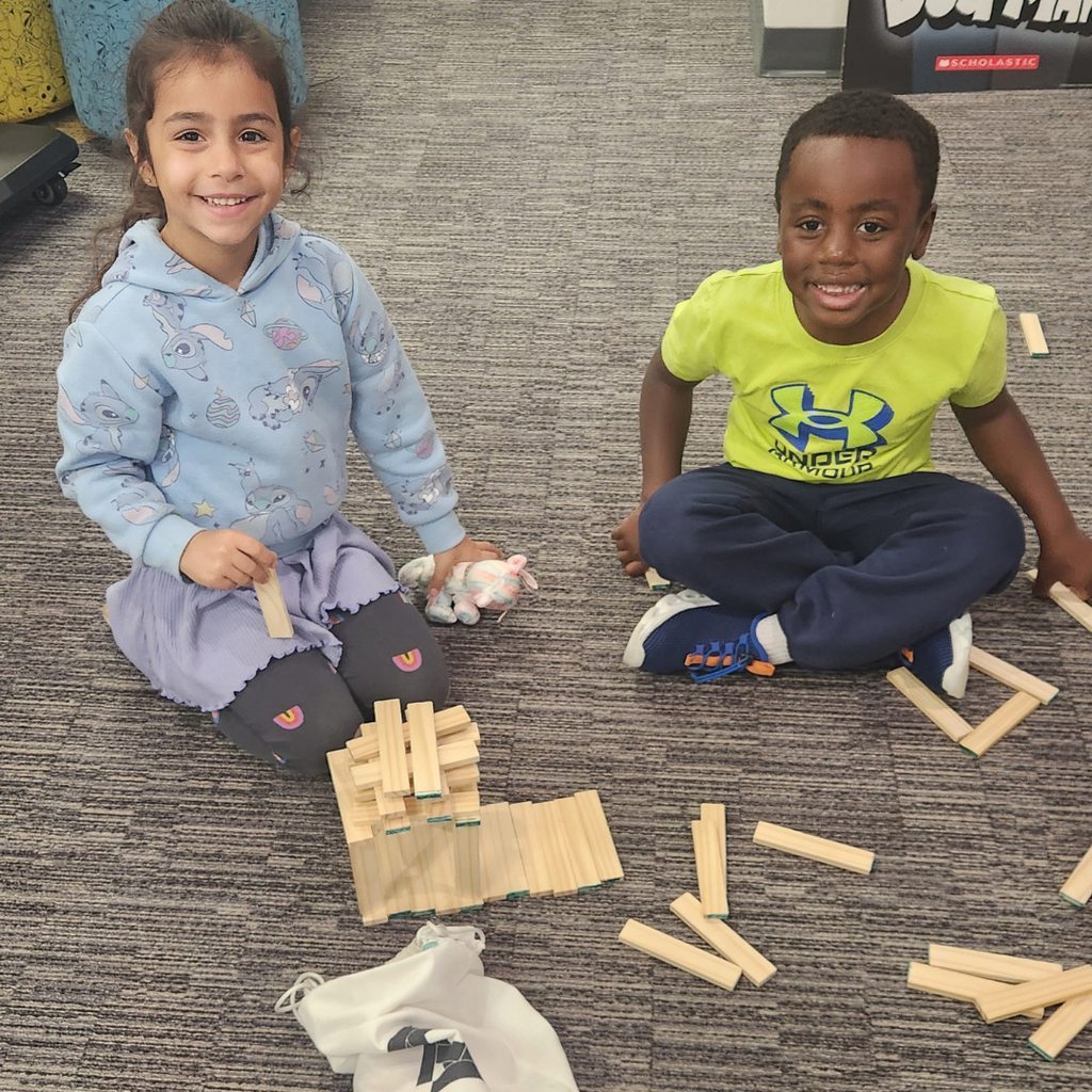 Two students smile next to library bookshelves as they build small wooden plank towers on the carpet.
