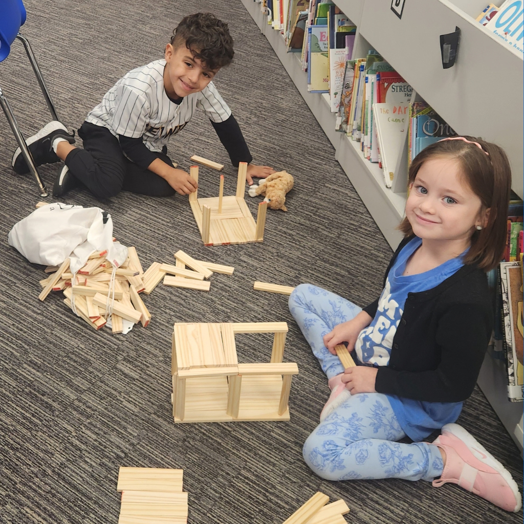 Two smiling elementary students sit on the floor in a library while constructing shapes out of wooden planks.