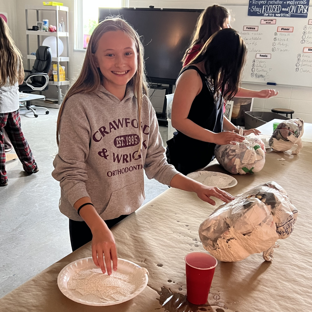 A smiling student in a gray sweatshirt participating in a paper mâché art project at a long table in a classroom.