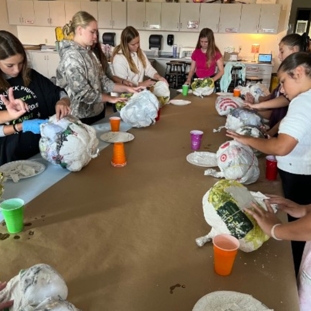 Students gathered around a long table covered in kraft paper, working on papier-mâché sculptures during an art class.