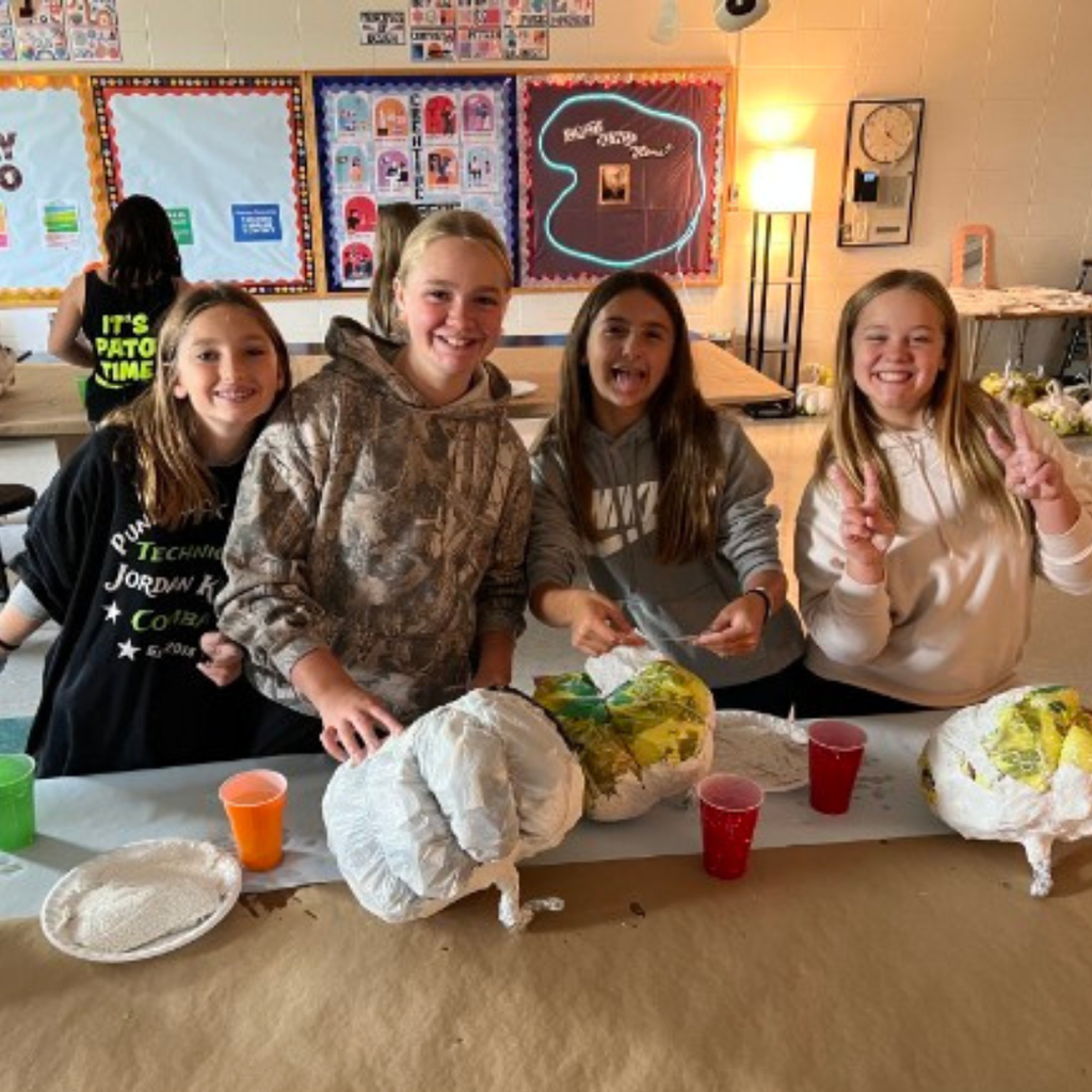 Four smiling students posing together behind a table displaying their papier-mâché art projects in a classroom.