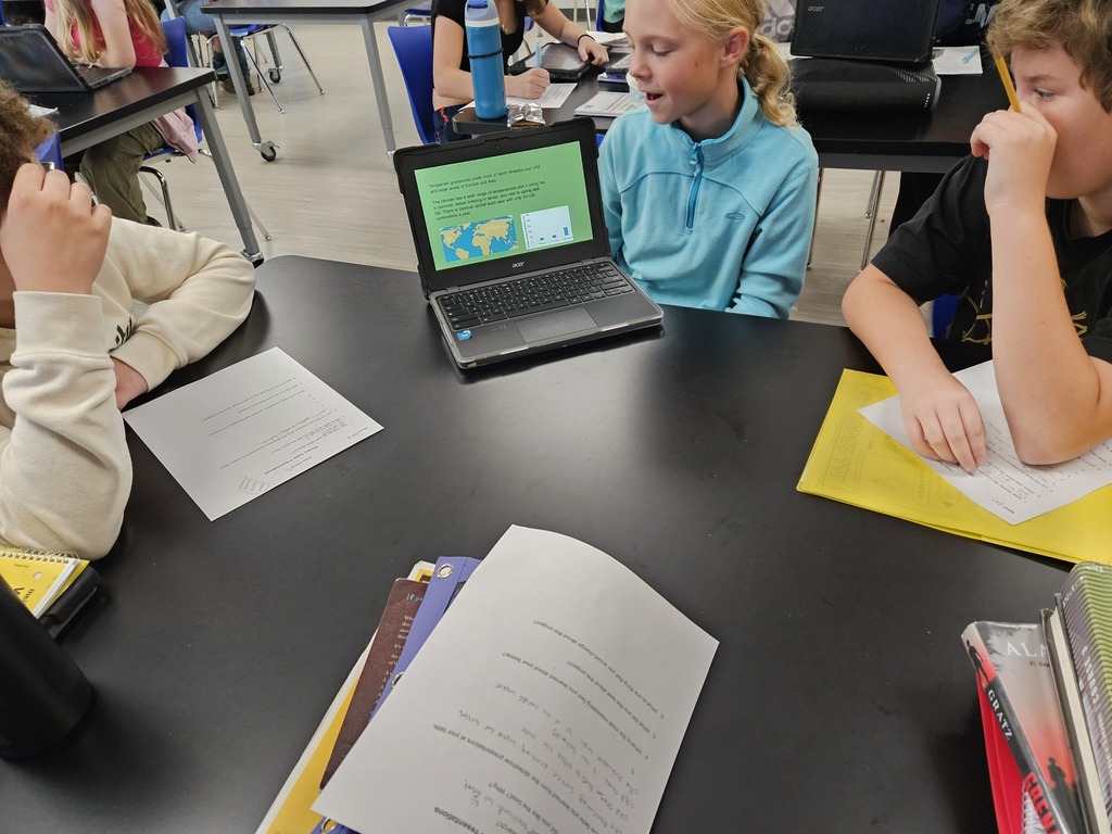 Students sitting around a classroom table reviewing a slide presentation on a laptop together.