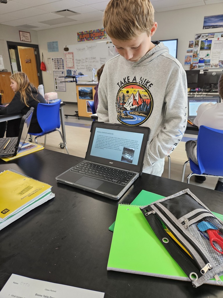 A student in a gray hoodie standing at a classroom desk looking down at a presentation open on a laptop.