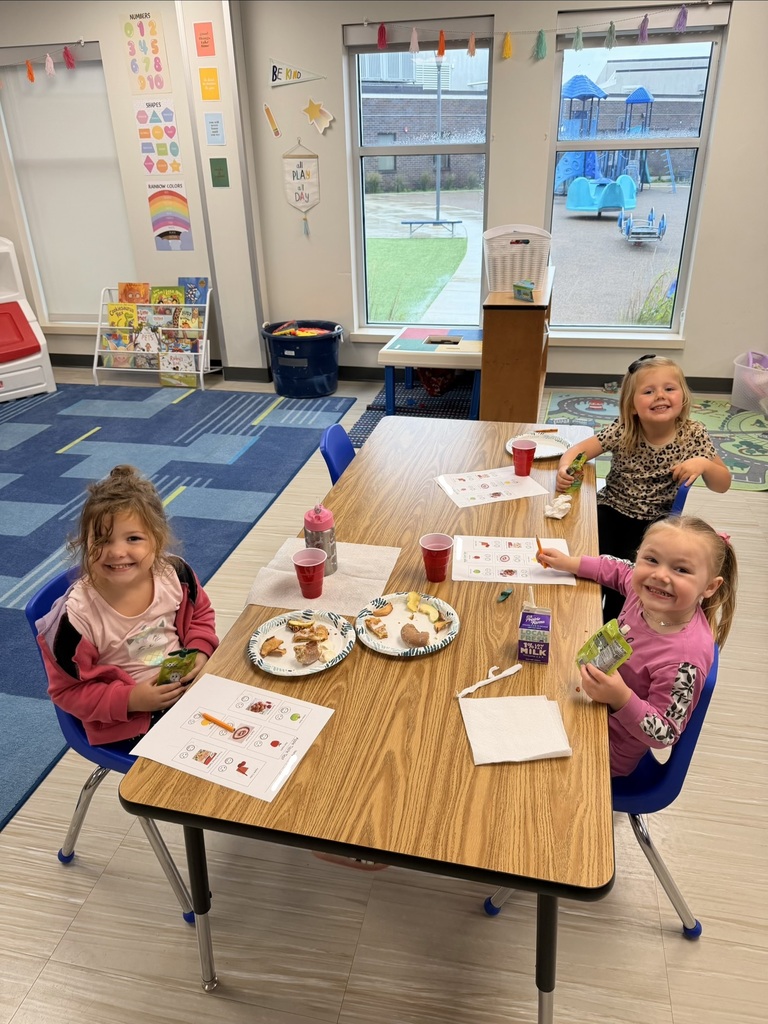 Three young students seated at a classroom table smiling near windows facing a playground.