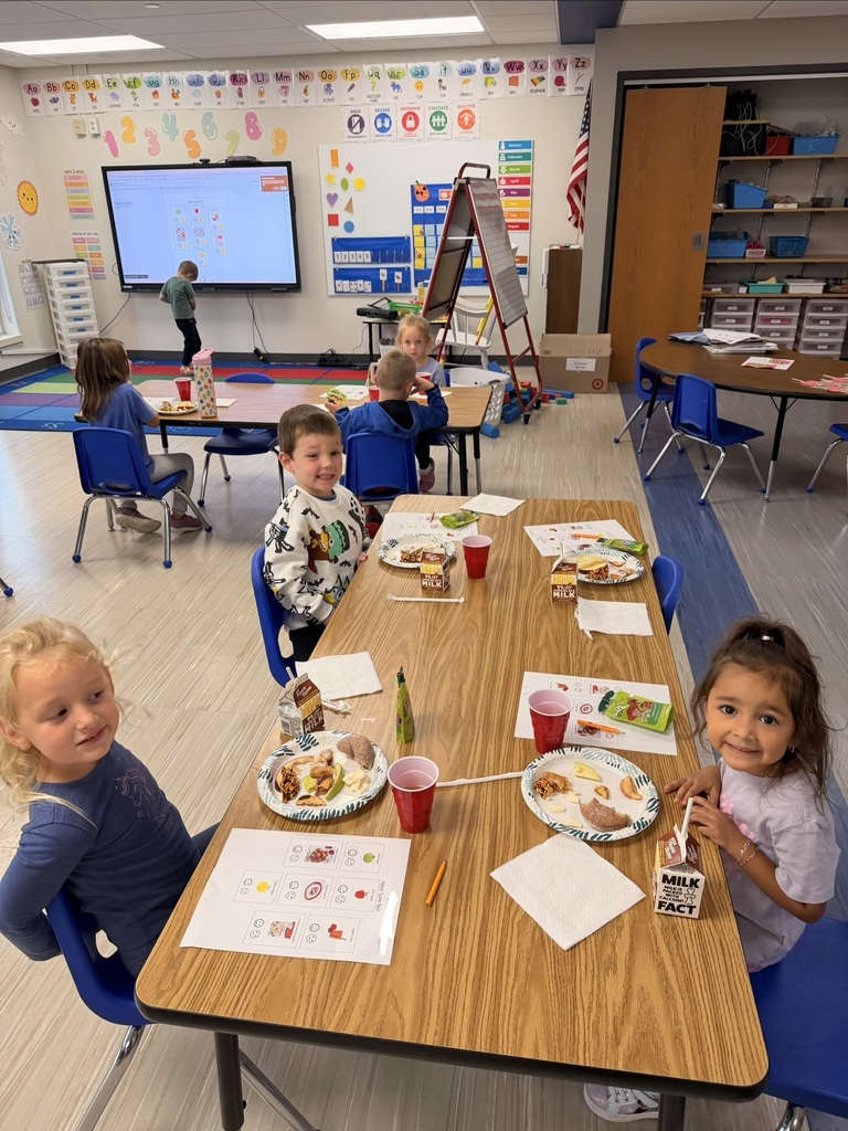 Elementary students sitting at tables with paper plates, drinks, and worksheets during class snack time.