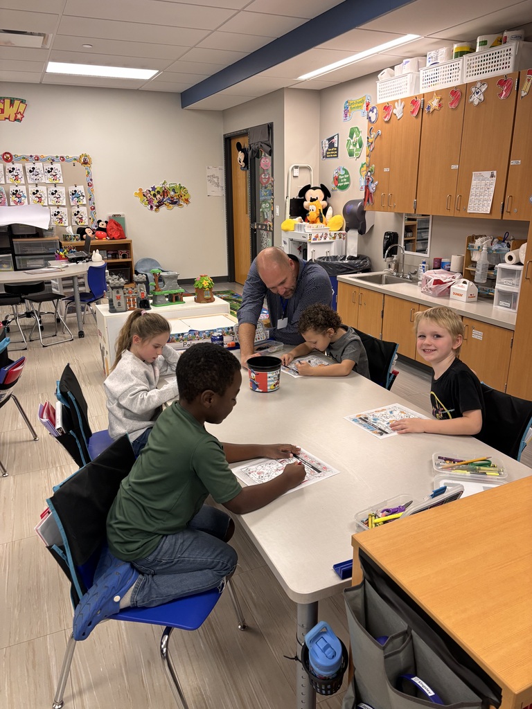 Four young students color activity sheets at a classroom table while a Mr. McBurney leans in to help a student in a bright elementary classroom.