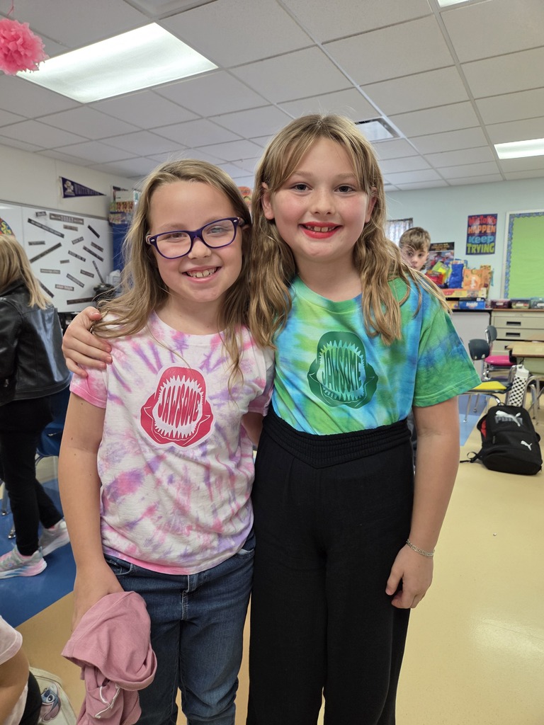 Two young elementary school friends smiling together while wearing matching tie-dye shark t-shirts.