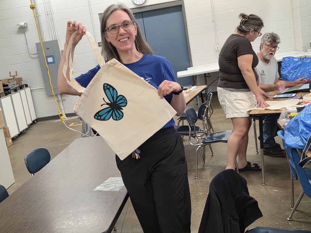 A smiling woman wearing glasses holding up a completed canvas tote bag featuring a blue monarch butterfly design.