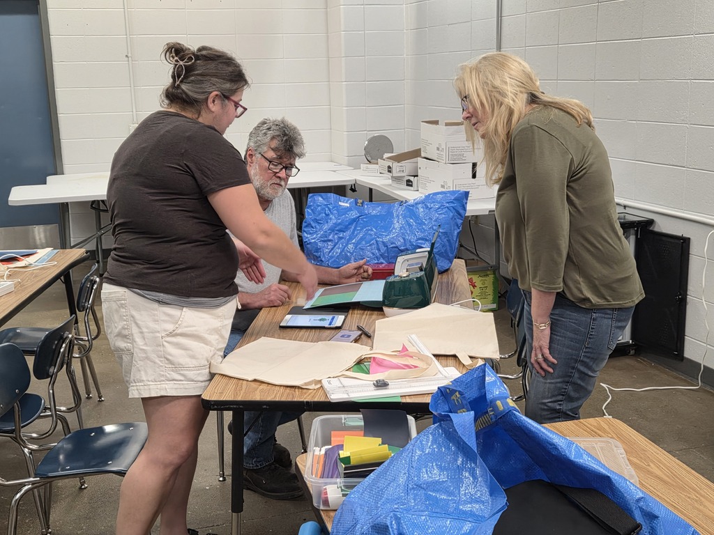 Three adults collaborating around a table with a tablet, crafting materials, and vinyl cutting machine in a school workshop room.