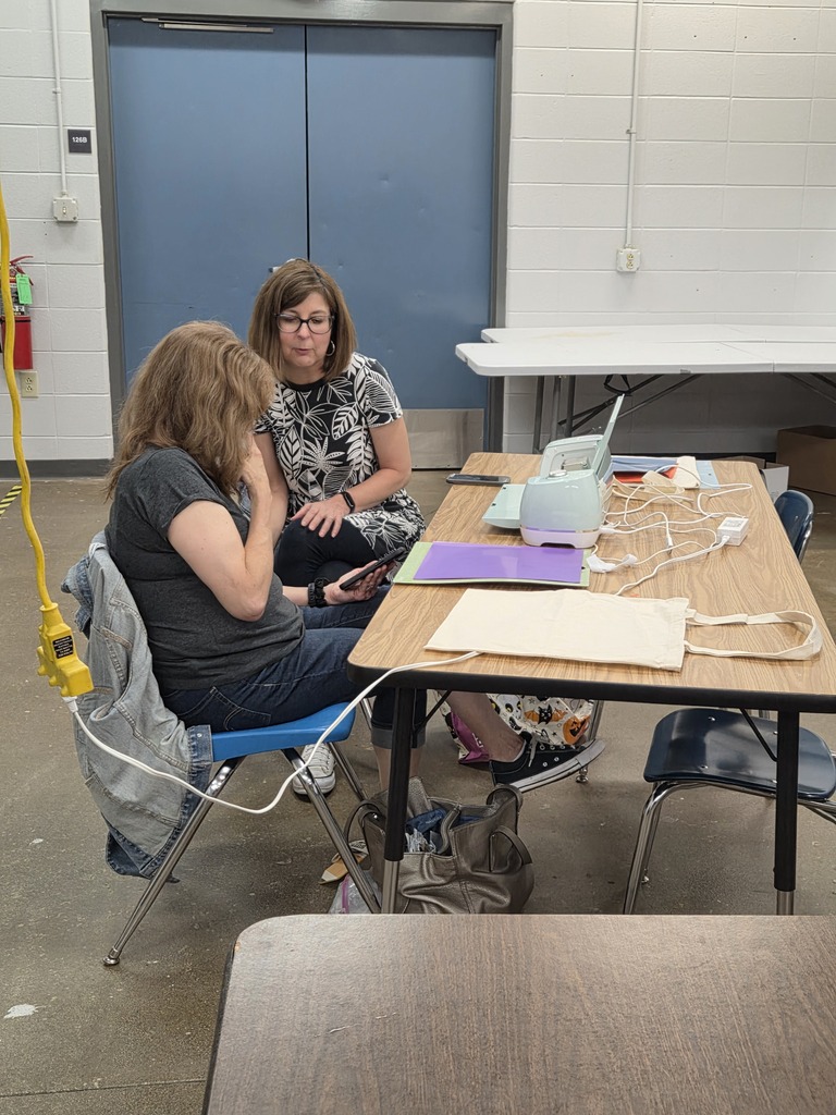 Two women seated together at a worktable looking at a smartphone next to a vinyl cutting machine and canvas tote bag.
