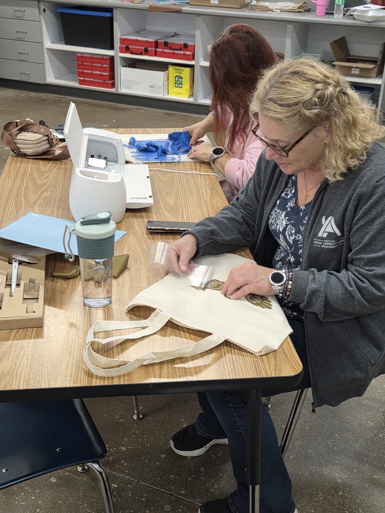 A woman applying a vinyl heat-transfer decal to a custom canvas tote bag at a table with cutting machines and craft supplies.