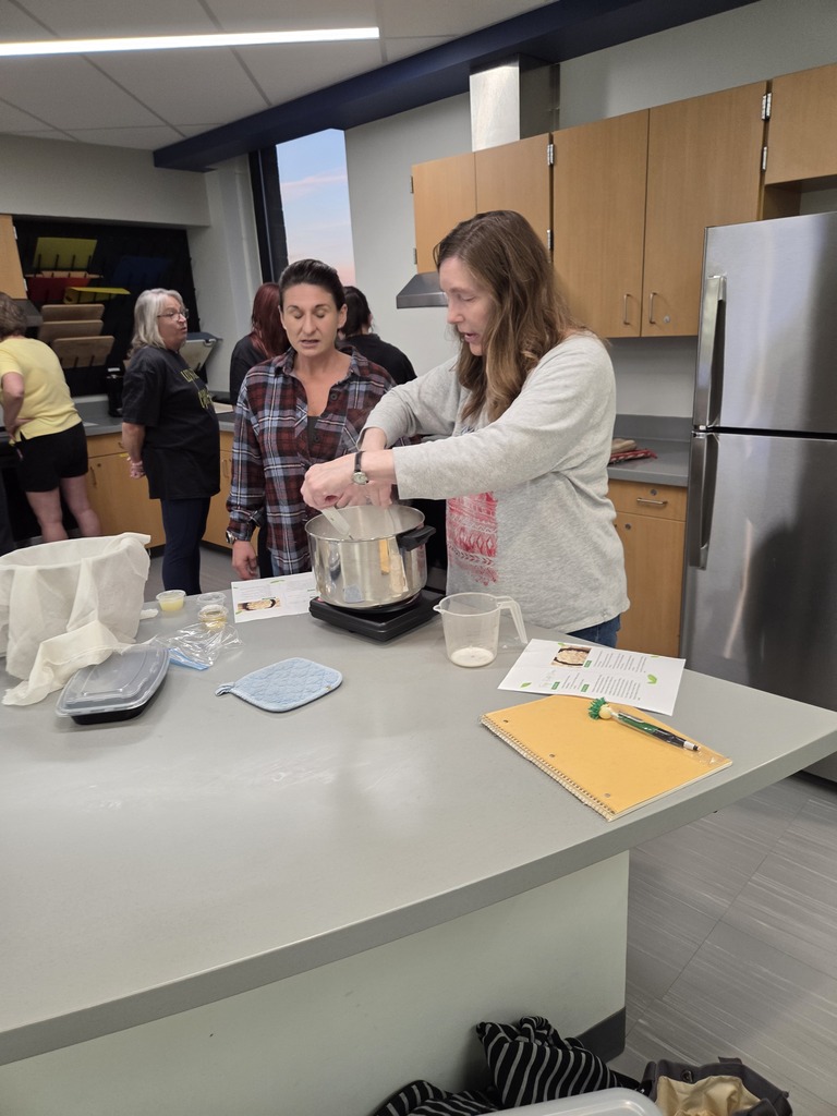 A woman demonstrating a cooking technique to a student watching closely in a school kitchen lab setting.