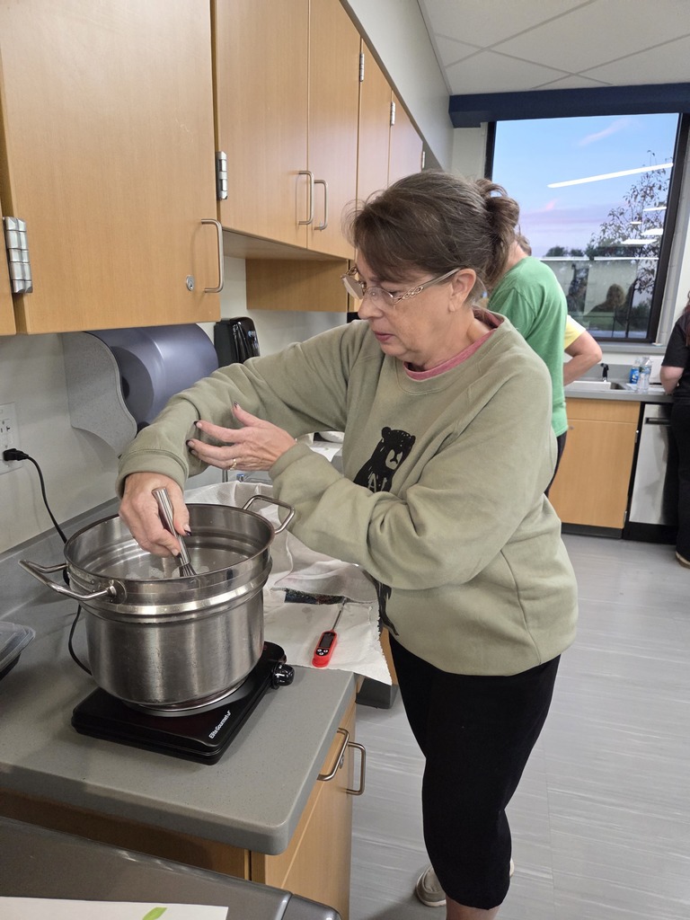 A woman stirring a metal pot on a hot plate during a cooking workshop in a school classroom kitchen.