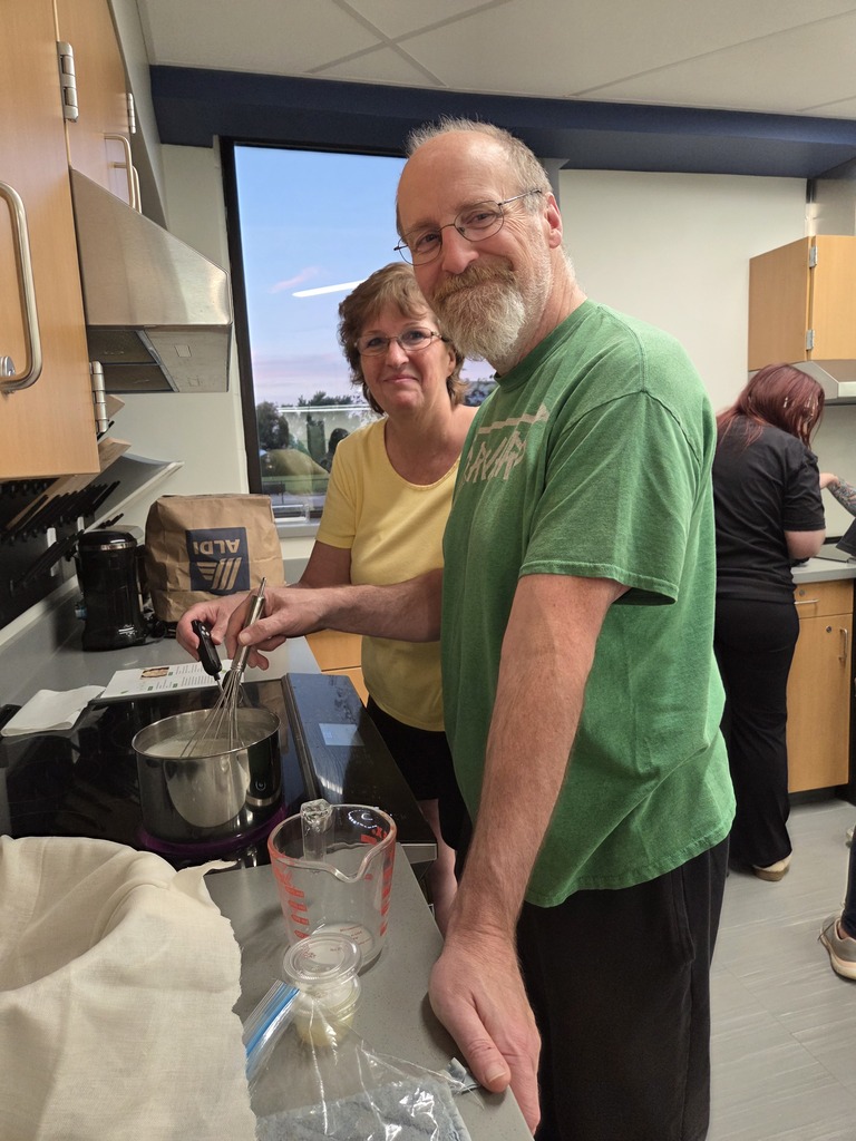 A man with a whisk stirring a pot while smiling next to a woman in a classroom kitchen setting.