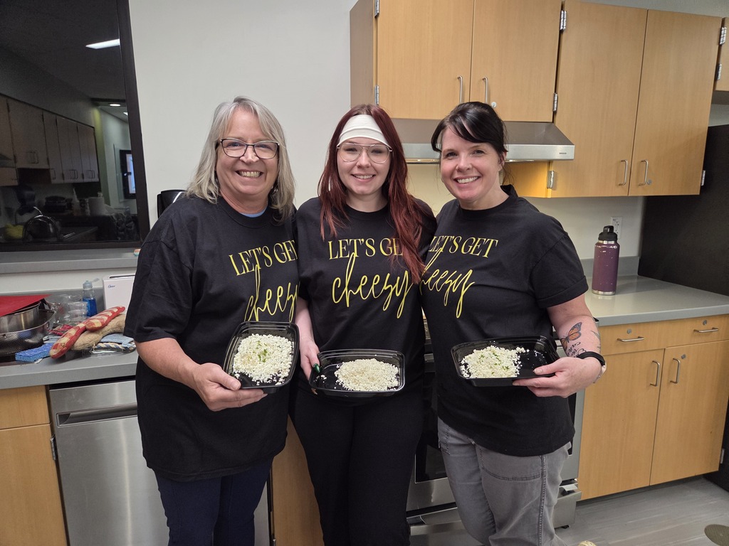 Three women wearing matching "Let's Get Cheesy" shirts smiling while holding trays of prepared food in a school kitchen.
