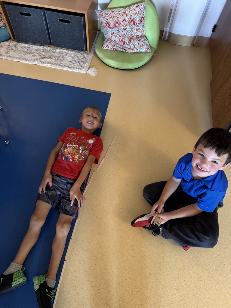 Two young boys smiling on the classroom floor, one lying on a blue mat and the other sitting cross-legged.