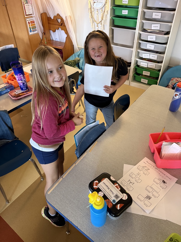Two smiling elementary girls standing near a classroom worktable, with one girl holding up a piece of paper.