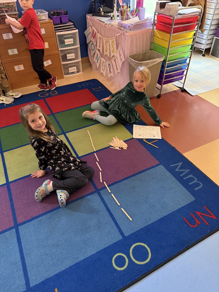 Two young students sitting on an alphabet classroom rug, using popsicle sticks to complete a math activity.