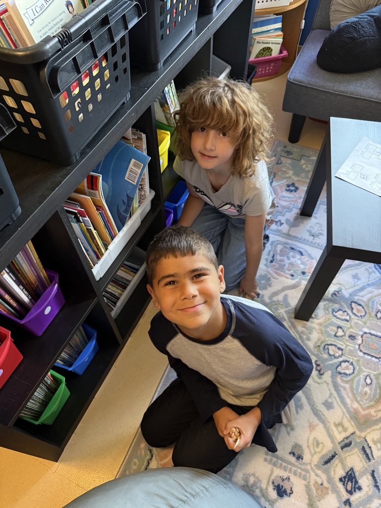 Two elementary students kneeling on the floor near a classroom bookshelf, smiling together during learning time.