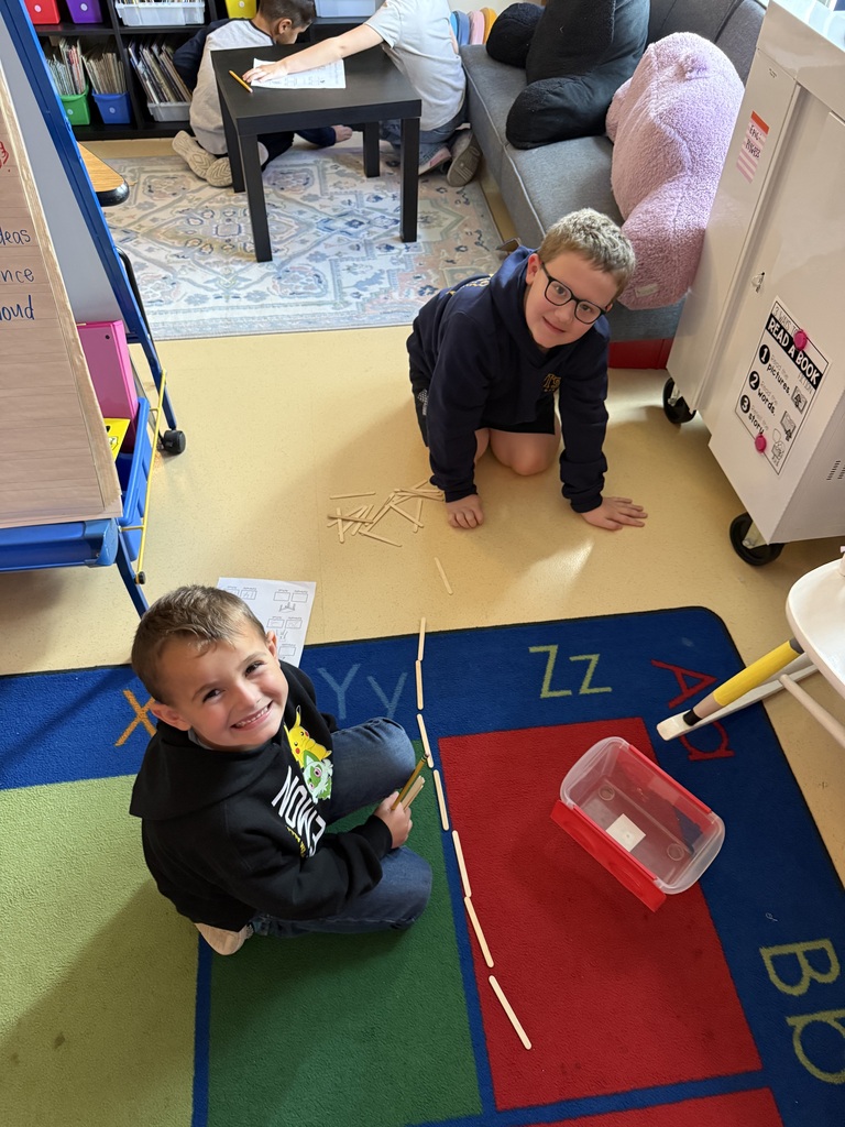 Two young boys sitting on the classroom floor smiling while working on a measurement activity with craft sticks