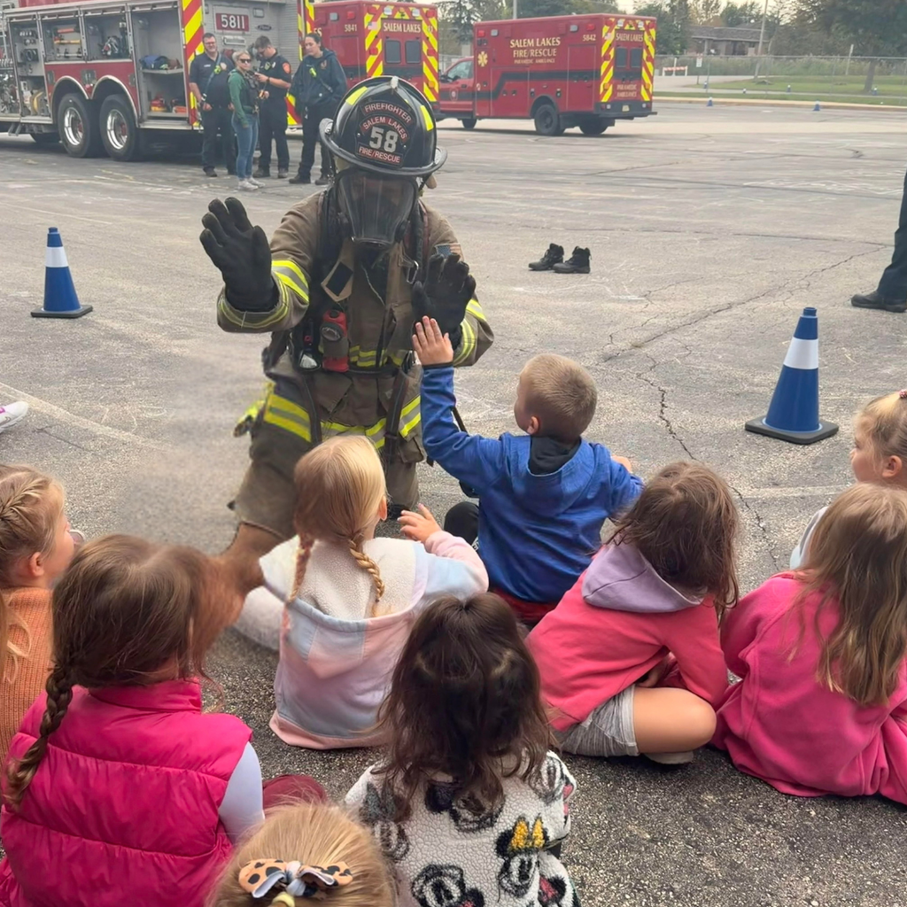 Elementary students sitting outside and high-fiving a firefighter in full turn-out gear during a Salem Lakes Fire Rescue visit.