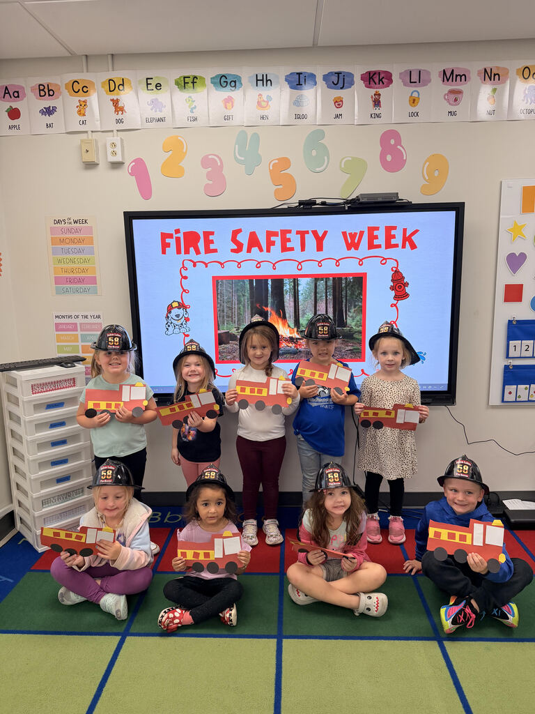 Students wearing toy firefighter helmets and holding paper fire truck crafts in front of a classroom display for Fire Safety Week.