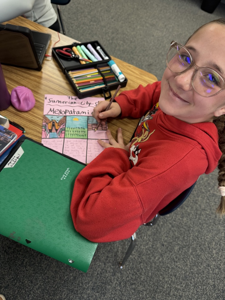 A student smiling at her desk while working on an illustrated poster project about Sumerian city-states in Mesopotamia.