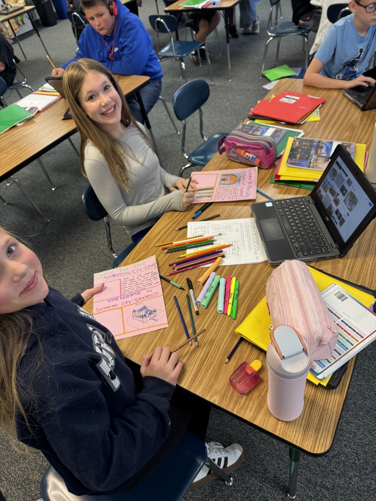 Students smiling at their desks in a classroom surrounded by art supplies and posters about Sumerian city-states.