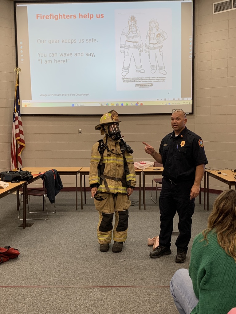 A firefighter in full gear stands beside a fire official giving a fire safety presentation to students in front of a projection screen.