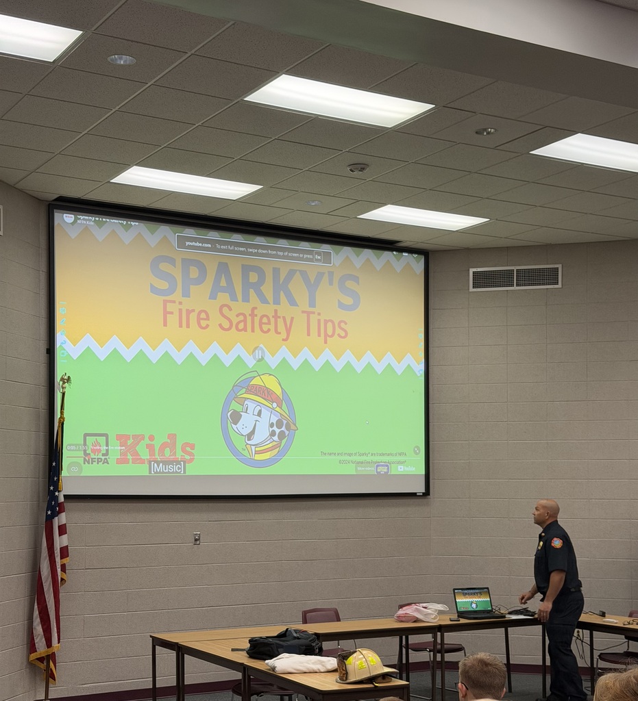 A firefighter stands next to a large projection screen displaying "Sparky's Fire Safety Tips" during a school assembly.