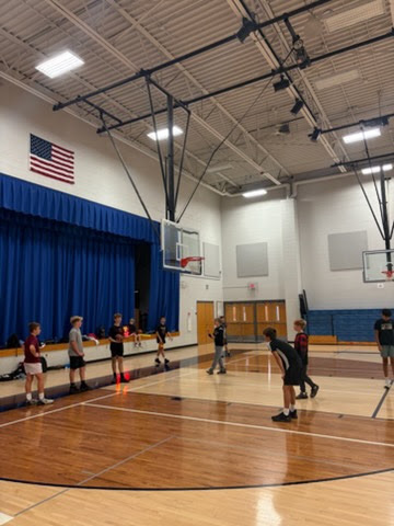 Students participating in open gym on a basketball court in front of a stage with blue curtains.