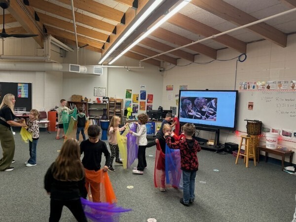 Elementary students waving colorful scarves while following along with a video on a large digital display.