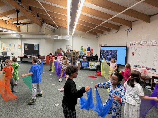 Elementary students wearing pajamas and dancing with colorful sheer scarves in a music classroom.