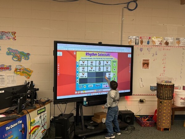 A student touching a large interactive display screen during a digital music rhythm activity.