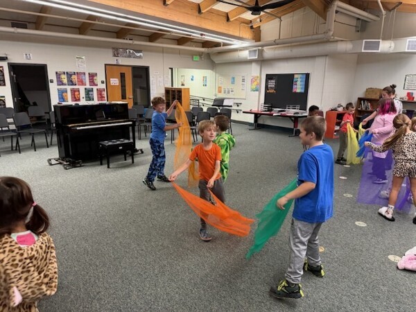 Elementary students dancing and waving colorful scarves during a music class with an upright piano in the background.