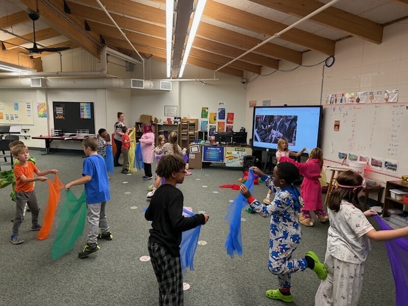 Young students in pajamas practicing movement and rhythm with colorful scarves during music class.