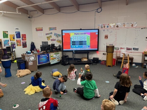Students sitting on the classroom floor holding musical shakers during an interactive display lesson.