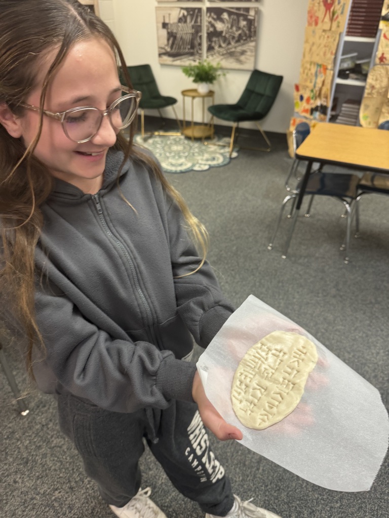 A student holding up a completed cuneiform clay tablet on wax paper in a classroom.